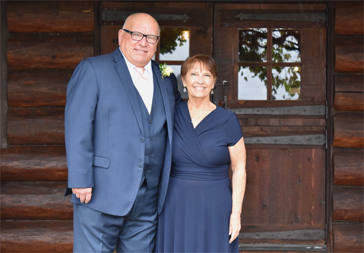 David Haire and wife Anna smiling and posing for a picture in front of a log cabin with a heavy wood door.